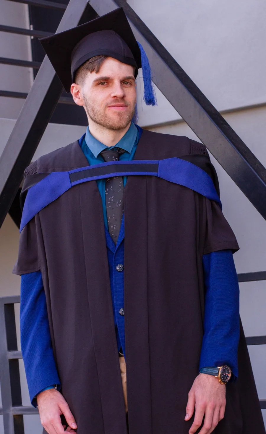 Ruan Leenders in a graduation gown, cap and blue hood, standing on a black staircase